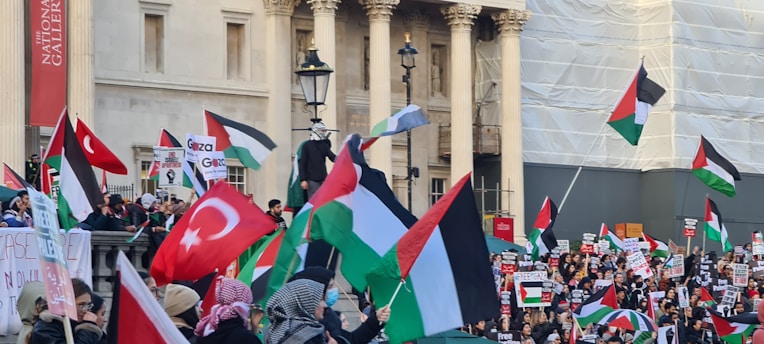 A large crowd of people is gathered outside a building with columns, holding flags and signs. The flags include Turkish and Palestinian national flags. The participants appear to be engaged in a demonstration or protest.