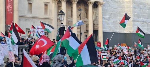 A large crowd of people is gathered outside a building with columns, holding flags and signs. The flags include Turkish and Palestinian national flags. The participants appear to be engaged in a demonstration or protest.