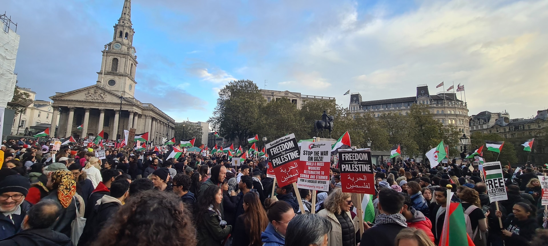 Wide angle shot of Tahrir Square filled with thousands of demonstrators holding banners and flags, under a dramatic cloudy sky.