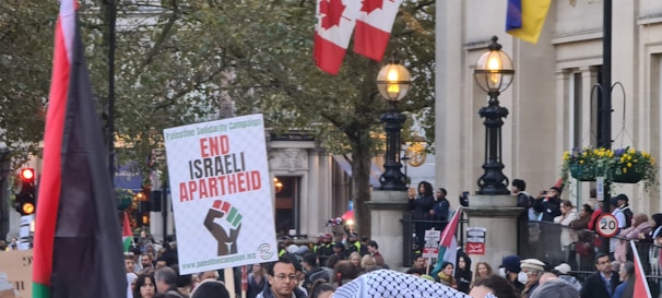 A protest scene with a crowd of people holding signs and flags. One prominent sign reads 'End Israeli Apartheid' with a graphic of a clenched fist. The background shows trees, streetlights, and a building adorned with various flags, including the Canadian flag.
