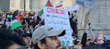A large crowd of people gathered in a public space, holding signs and flags. Many of the signs contain political messages, and several flags are visible, including those with red, black, green, and white colors. The scene appears to be a protest or demonstration with diverse participants engaged in activism.
