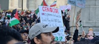 A large crowd of people gathered in a public space, holding signs and flags. Many of the signs contain political messages, and several flags are visible, including those with red, black, green, and white colors. The scene appears to be a protest or demonstration with diverse participants engaged in activism.