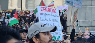 A large crowd of people gathered in a public space, holding signs and flags. Many of the signs contain political messages, and several flags are visible, including those with red, black, green, and white colors. The scene appears to be a protest or demonstration with diverse participants engaged in activism.
