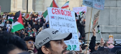 A large crowd of people gathered in a public space, holding signs and flags. Many of the signs contain political messages, and several flags are visible, including those with red, black, green, and white colors. The scene appears to be a protest or demonstration with diverse participants engaged in activism.
