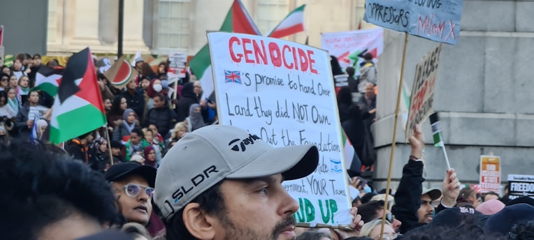 A large crowd of people gathered in a public space, holding signs and flags. Many of the signs contain political messages, and several flags are visible, including those with red, black, green, and white colors. The scene appears to be a protest or demonstration with diverse participants engaged in activism.