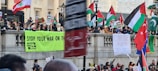 A large group of people gathered at a public demonstration, holding signs and banners with messages such as 'Stop Your War on the Climate' and 'Ceasefire Now'. Various national flags are visible, including those of Palestine, Lebanon, and Pakistan. The crowd stands behind a stone railing in an urban setting.