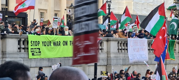 A large group of people gathered at a public demonstration, holding signs and banners with messages such as 'Stop Your War on the Climate' and 'Ceasefire Now'. Various national flags are visible, including those of Palestine, Lebanon, and Pakistan. The crowd stands behind a stone railing in an urban setting.
