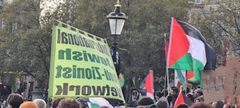 A group of people gathered outdoors, holding flags and signs. The flags display red, black, white, and green colors, and one of the signs has bold text with a green background. Trees and a streetlight are visible in the background.