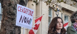 A young person with long hair stands in front of a building with Canadian flags. Next to them, a tree has a sign attached that reads 'Ceasefire Now!' in bold letters.