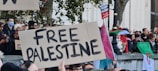A group of people participating in a demonstration, holding signs and flags. The prominent sign reads 'FREE PALESTINE' in bold letters. Many individuals are visible, some wearing masks, with various banners and flags, including a Palestinian flag. The scene is set outdoors with trees and buildings in the background.