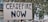A cardboard sign with the bold black letters 'CEASEFIRE NOW' held in an outdoor environment. Behind the sign, a glimpse of a tree with green leaves and a portion of a building is visible. Additional signs can be partially seen in the background.