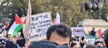 A large crowd is gathered for a protest with people holding signs and waving flags. The signs display messages about stopping ethnic cleansing and supporting Gaza. In the background, there is a large equestrian statue.