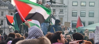 A large crowd of people gathered with various flags, predominantly displaying red, black, white, and green colors. Many participants hold signs, some of which call for ceasefire and express political messages. The scene takes place in an urban environment, with buildings and windows visible in the background.
