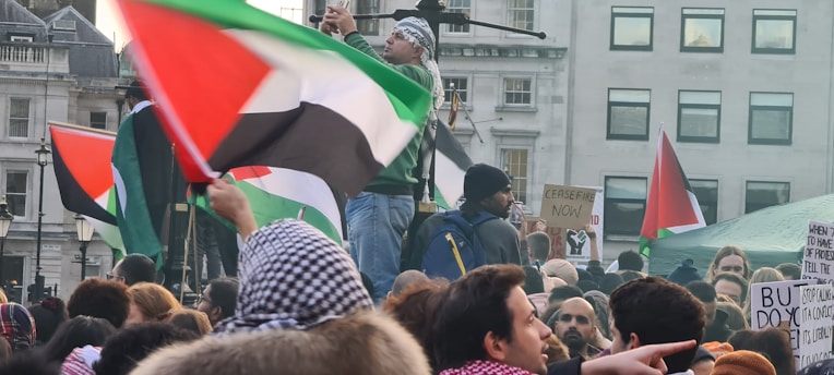 A large crowd of people gathered with various flags, predominantly displaying red, black, white, and green colors. Many participants hold signs, some of which call for ceasefire and express political messages. The scene takes place in an urban environment, with buildings and windows visible in the background.