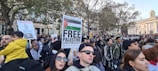 A large crowd gathers for a protest, with people holding various signs advocating for Palestinian rights. The surroundings include trees and a stately building in the background. Prominent signs include ones that read 'Free Palestine' and 'Ceasefire Now.' The crowd is diverse, with individuals wearing a mix of casual and cultural attire.