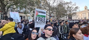 A large crowd gathers for a protest, with people holding various signs advocating for Palestinian rights. The surroundings include trees and a stately building in the background. Prominent signs include ones that read 'Free Palestine' and 'Ceasefire Now.' The crowd is diverse, with individuals wearing a mix of casual and cultural attire.