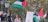 A group of people, some holding Palestinian flags, are gathered in an area with street lamps and a statue in the background. The flags are prominently displayed, and a couple of individuals are seen using their phones. There are trees in the background, and signs with text are partially visible.