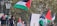 A group of people, some holding Palestinian flags, are gathered in an area with street lamps and a statue in the background. The flags are prominently displayed, and a couple of individuals are seen using their phones. There are trees in the background, and signs with text are partially visible.