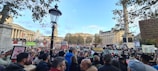 A large crowd is gathered in an urban square, holding various signs and banners with messages related to political issues. In the background, there are historic buildings, a column, and trees. The weather appears clear with blue sky and scattered clouds.