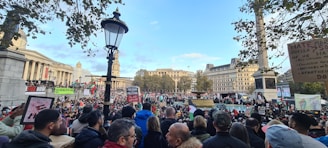 A large crowd is gathered in an urban square, holding various signs and banners with messages related to political issues. In the background, there are historic buildings, a column, and trees. The weather appears clear with blue sky and scattered clouds.