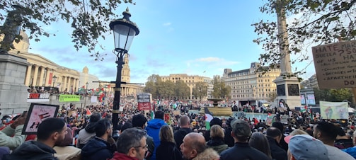 A large crowd is gathered in an urban square, holding various signs and banners with messages related to political issues. In the background, there are historic buildings, a column, and trees. The weather appears clear with blue sky and scattered clouds.