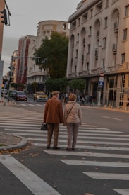 An elderly couple walks hand in hand across a city street at a crosswalk. The street is lined with tall buildings, including a prominent one with a 'Gucci' sign. The autumnal leaves are scattered on the ground, and a few people and cars are seen in the background.