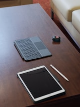 A neatly arranged workspace on a wooden table featuring a tablet with a dark screen accompanied by a stylus. Adjacent to the tablet, there's a detachable keyboard case. A small black remote control with a red button is also present on the table. In the background, parts of two cream-colored sofas are visible.