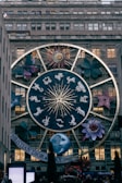 A large zodiac-themed structure is displayed on the facade of a building, featuring a prominent circular design with the twelve zodiac signs, a stylized sun with a face, and various large floral and cosmic elements around it. The scene is in a city setting with a specific brand name visible at the bottom.