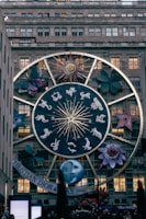 A large zodiac-themed structure is displayed on the facade of a building, featuring a prominent circular design with the twelve zodiac signs, a stylized sun with a face, and various large floral and cosmic elements around it. The scene is in a city setting with a specific brand name visible at the bottom.
