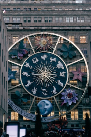A large zodiac-themed structure is displayed on the facade of a building, featuring a prominent circular design with the twelve zodiac signs, a stylized sun with a face, and various large floral and cosmic elements around it. The scene is in a city setting with a specific brand name visible at the bottom.