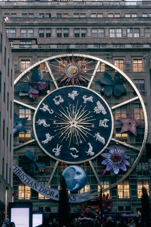 A large zodiac-themed structure is displayed on the facade of a building, featuring a prominent circular design with the twelve zodiac signs, a stylized sun with a face, and various large floral and cosmic elements around it. The scene is in a city setting with a specific brand name visible at the bottom.
