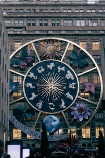 A large zodiac-themed structure is displayed on the facade of a building, featuring a prominent circular design with the twelve zodiac signs, a stylized sun with a face, and various large floral and cosmic elements around it. The scene is in a city setting with a specific brand name visible at the bottom.