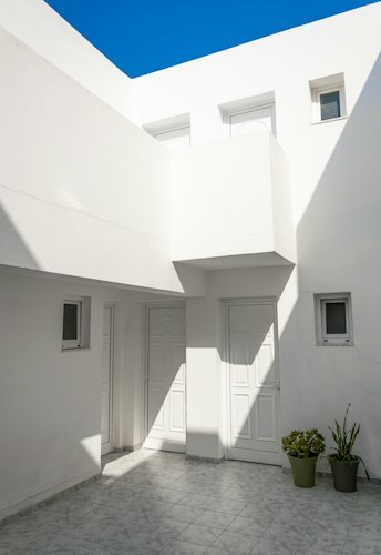A minimalist courtyard is surrounded by white walls and features clean architectural lines. The area is enclosed with multiple white doors and small rectangular windows. The shadow play on the walls and the floor adds depth to the space. Two potted plants are placed at the corner, adding a touch of greenery. The bright blue sky above contrasts with the stark white of the architecture.