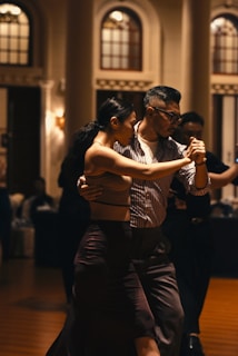 A couple elegantly dances in a dimly lit room with an ambiance created by warm lighting and classic architecture. Their coordinated movements suggest they are engaged in a formal dance, possibly a tango, with focused expressions and close physical proximity.