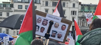A protest scene with people holding signs and flags. A central sign labeled 'Faces of Evil' displays multiple faces. Various posters and banners with political messages are visible, and the setting is likely an urban environment with historical architecture.