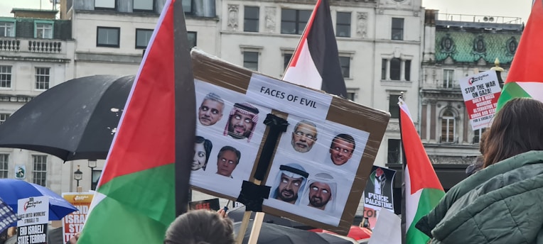A protest scene with people holding signs and flags. A central sign labeled 'Faces of Evil' displays multiple faces. Various posters and banners with political messages are visible, and the setting is likely an urban environment with historical architecture.