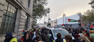 A large group of people gathered in a city street for a demonstration. Many individuals are carrying flags with red, green, black, and white colors. The scene is energetic, with a police vehicle present amidst the crowd. The setting is urban, with historical buildings and a cloudy sky overhead.