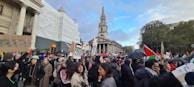 A large crowd of people gathered in a public square, holding signs and flags. Many are wearing winter clothing, suggesting a cooler climate. In the background, there is an old building with columns and a church with a prominent spire. Some of the signs and flags are related to a political cause.