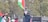 A group of people are seen holding flags with red, green, black, and white colors. One individual is prominently waving a large flag while another is capturing a photo or video on a phone. There are trees in the background and a decorative lamppost.