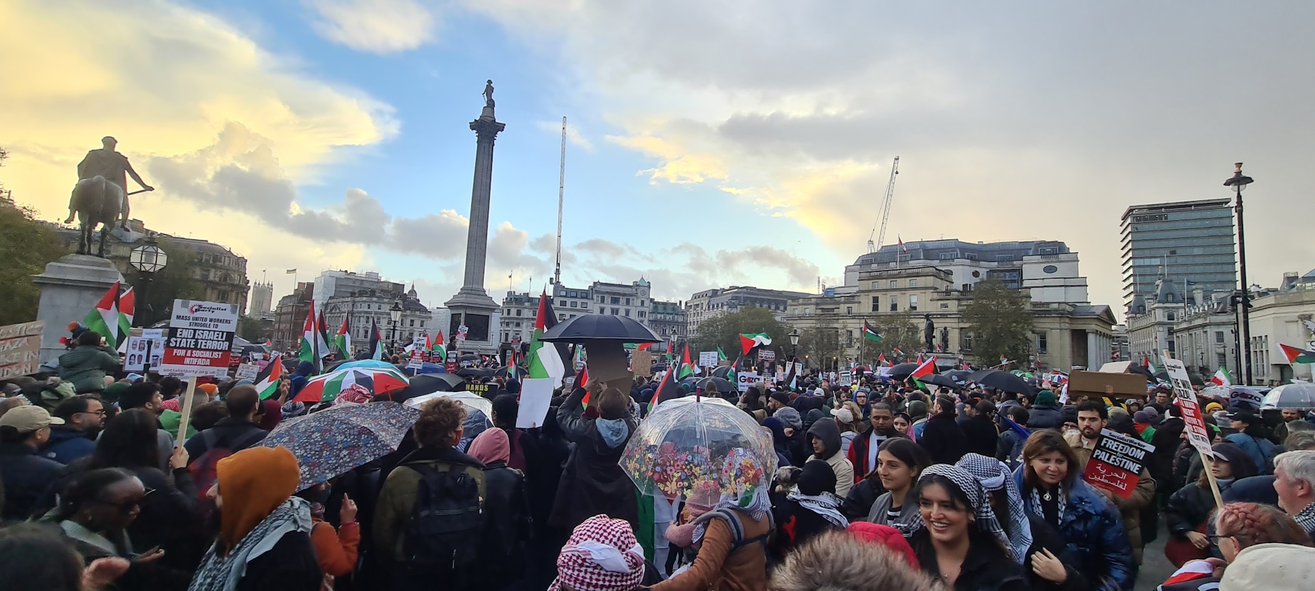 Wide angle shot of Tahrir Square filled with thousands of demonstrators holding banners and flags, under a dramatic cloudy sky.
