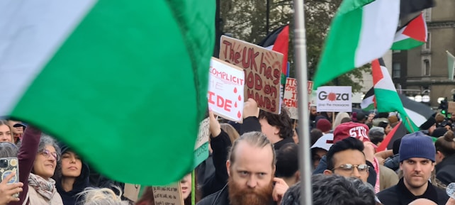 A large group of people gathers in a protest, holding up signs and flags. Several placards display messages about political issues. The Palestinian flag is prominently visible, waving in the air, and the crowd appears engaged and attentive.