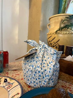 A floral-patterned fabric wrapped package sits on a table adorned with a flowered tablecloth. Nearby, a decorative ceramic plate features blue and red illustrations. In the background, a large ceramic pot with plant leaves and ornate script is visible, along with a red soda can and tissues.