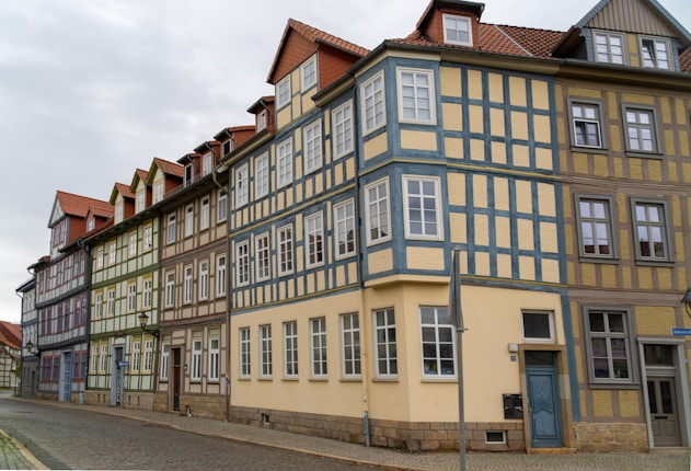 A row of colorful, traditional half-timbered houses with intricate wooden frameworks, situated along a cobblestone street. The buildings have multiple stories and are adorned with windows and steep gabled roofs. The overcast sky adds to the serene atmosphere of the scene.