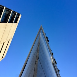An architectural photograph featuring a sharp, angular building structure pointing upward against a clear blue sky. Sunlight casts shadows, enhancing the building's clean lines and modern design. Another building with glass windows is visible on the left, adding to the urban landscape.