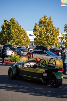 A green, open-roof, three-wheeled vehicle is driving in a parking lot. There are several people in the background, some walking and others standing near parked cars. Trees with green and yellow leaves are visible in the background, and the sky is clear and blue.