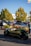 A green, open-roof, three-wheeled vehicle is driving in a parking lot. There are several people in the background, some walking and others standing near parked cars. Trees with green and yellow leaves are visible in the background, and the sky is clear and blue.