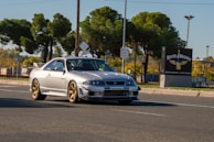 A silver sports car with a spoiler and aftermarket modifications is driving on a paved road. The background includes tall green trees, street signs, and a building with a sign that reads 'Rock & Brews' advertising a restaurant and live music.
