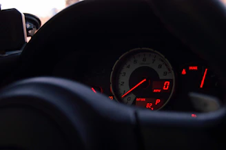 Close-up of a car dashboard showing the speedometer with a reading of zero. The instrument cluster features illuminated gauges with red and white backlights, and indicators for revolutions per minute, speed, and outside temperature. A partially visible steering wheel is in the foreground, and faint ambient light enters from the left.