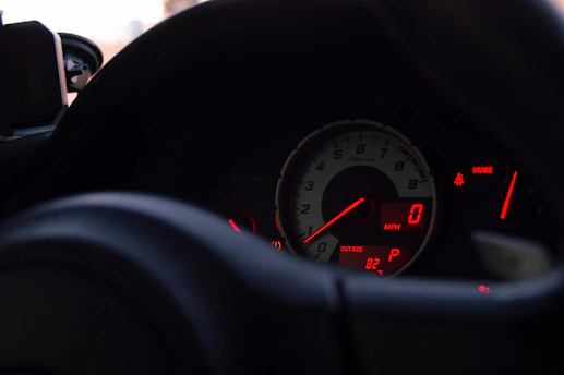 Close-up of a car dashboard showing the speedometer with a reading of zero. The instrument cluster features illuminated gauges with red and white backlights, and indicators for revolutions per minute, speed, and outside temperature. A partially visible steering wheel is in the foreground, and faint ambient light enters from the left.
