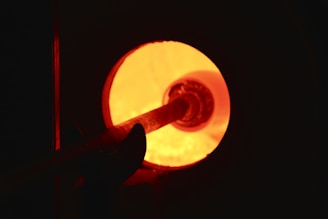 A glowing hot furnace with an intense orange light emanating from within. A metal tool is being inserted into the furnace, possibly for a glassblowing or metalworking process.